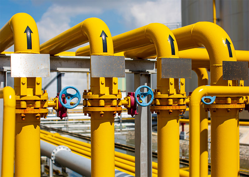 Yellow industrial gas pipes with black arrows indicating flow direction, connected by blue valve handles, set in an outdoor facility with additional pipes and a building in the background.