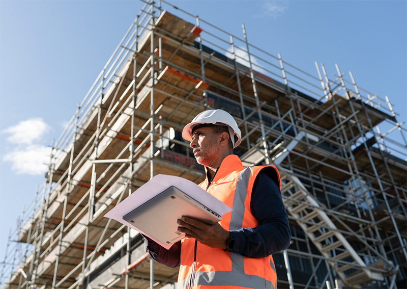 A construction worker wearing a white hard hat and orange safety vest holds documents while standing in front of a building under construction with visible scaffolding.