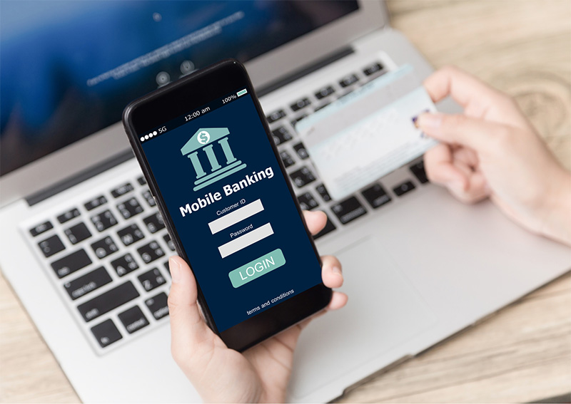 A person holds a smartphone displaying a mobile banking login screen, with a credit card in the other hand, in front of an open laptop on a wooden desk.