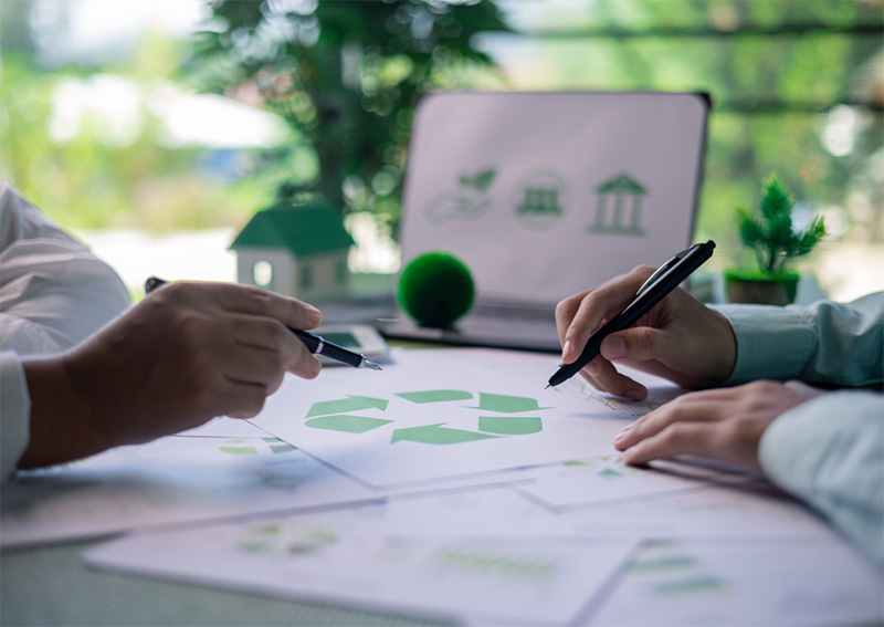 Two people discuss documents featuring a green recycling symbol. In the background are a laptop displaying eco-friendly icons, a small house model, a green ball, and potted plants, suggesting a focus on sustainability.