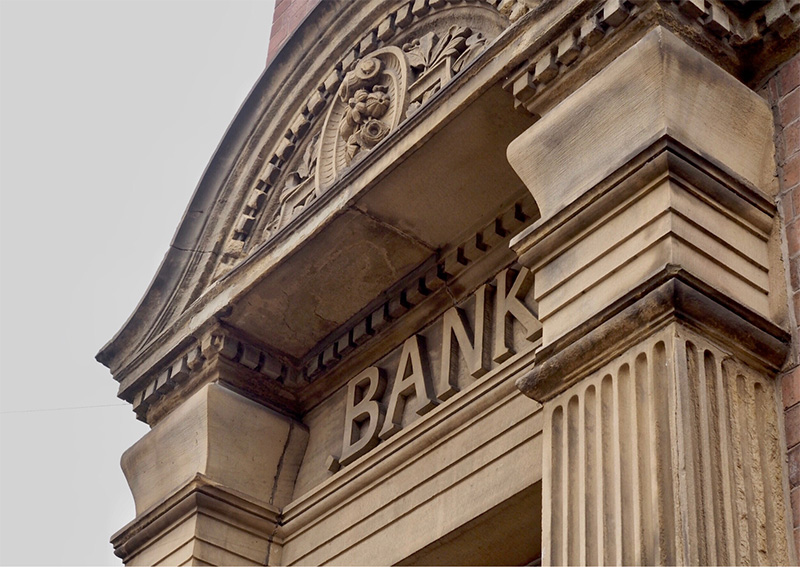 Close-up view of an old stone building with the word BANK engraved above the entrance, featuring ornate architectural details and columns.