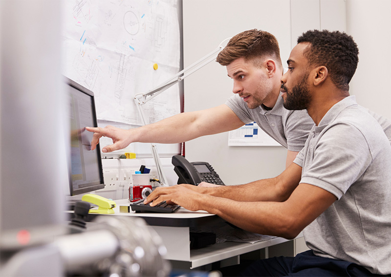 Two men in gray polo shirts work at a desk with a computer. One man is seated typing while the other stands, pointing at the screen. Technical drawings and office supplies are visible in the background.