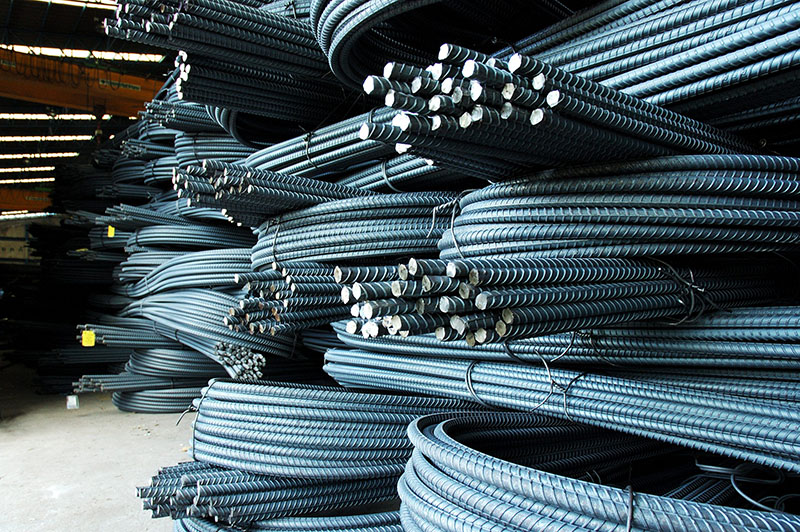 Stacks of bundled steel rebar rods are neatly arranged in a warehouse, with the ends of the rods facing outward and the coiled metal creating a pattern of lines and curves.