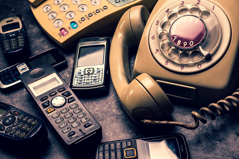 A collection of various phones, including a rotary dial telephone, cordless phones, and older mobile phones, displayed on a surface, showcasing the evolution of telephone technology.