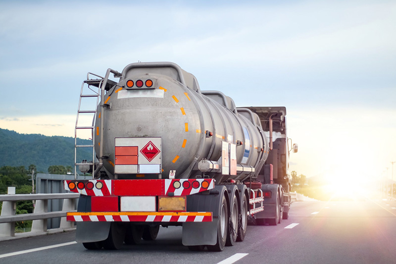 A large silver fuel tanker truck drives down a highway at sunset, with safety markings, hazard placards, and a ladder on the rear. The road curves ahead and a guardrail lines the side.