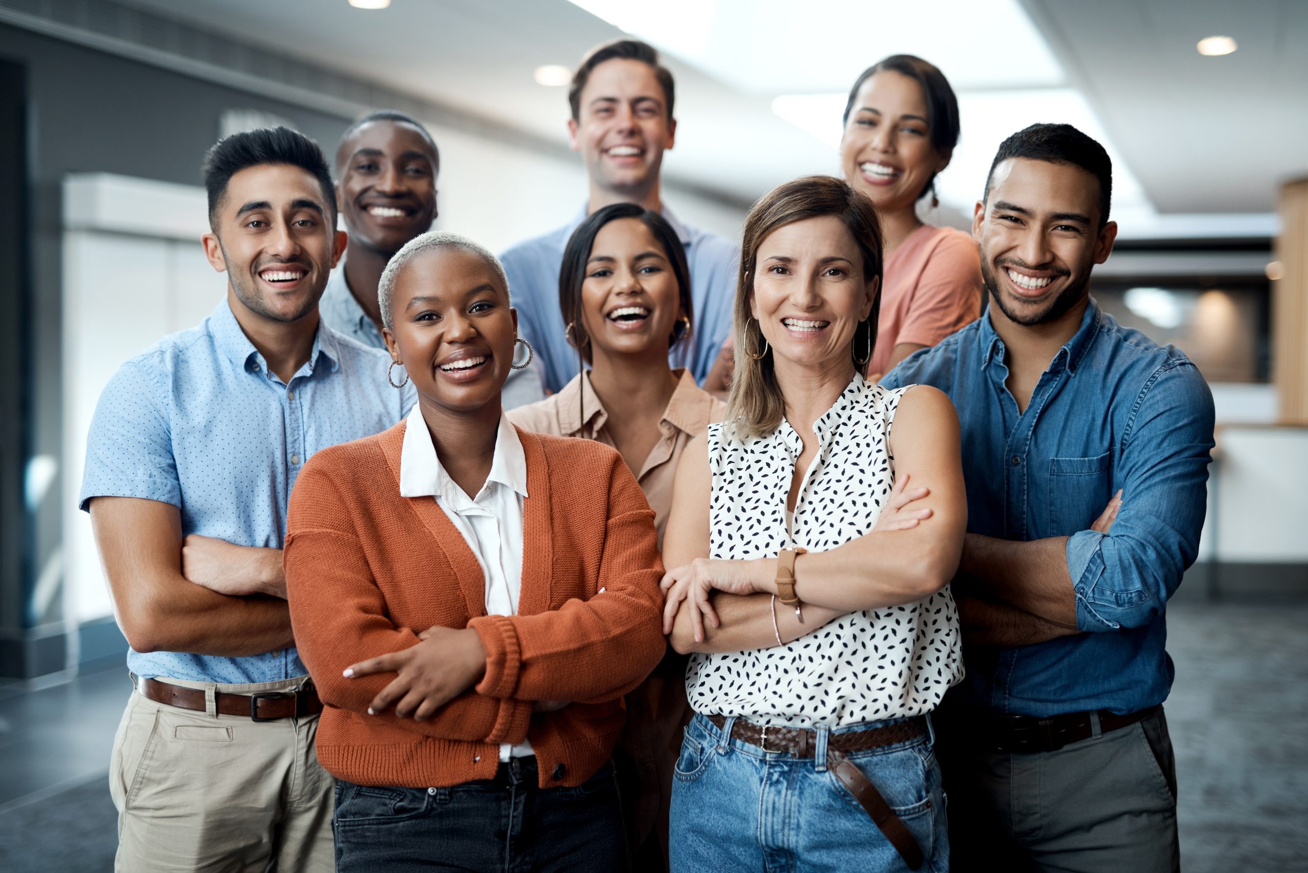 A diverse group of eight young adults stands close together indoors, smiling at the camera with arms crossed and appearing confident and friendly. The setting is a modern, well-lit building hallway.