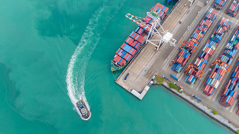 Aerial view of a cargo ship docked at a busy port with colorful shipping containers stacked on land and a small boat leaving a curved wake in turquoise water.