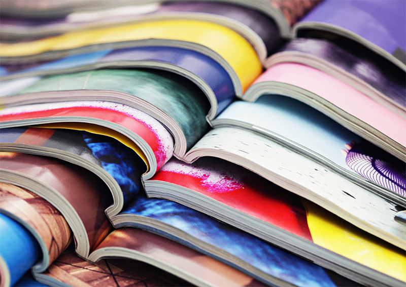 Close-up of several open magazines stacked on top of each other, showing colorful pages and curved spines, creating a layered and vibrant pattern.