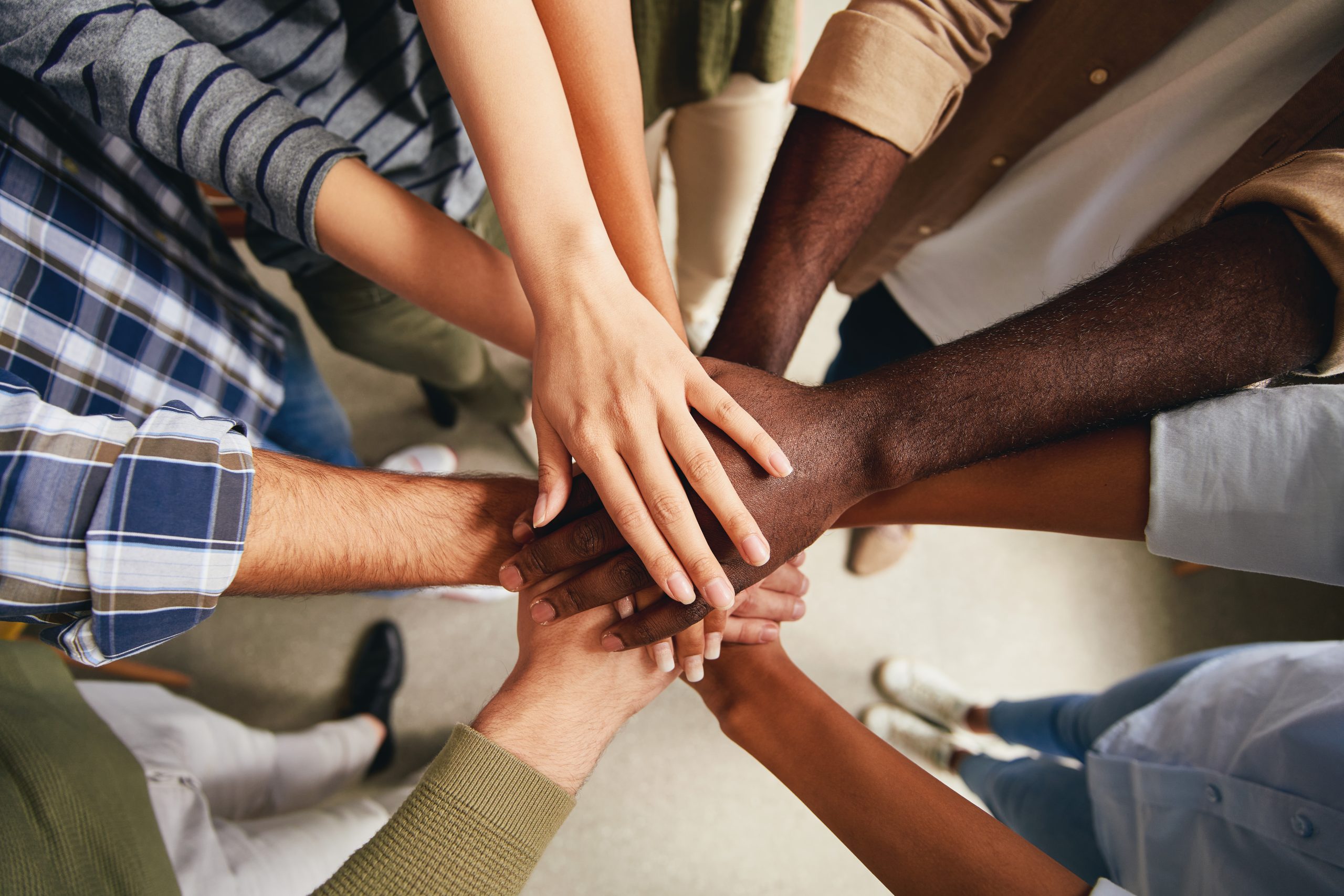 A diverse group of people stand in a circle and stack their hands together in the center, symbolizing teamwork and unity.