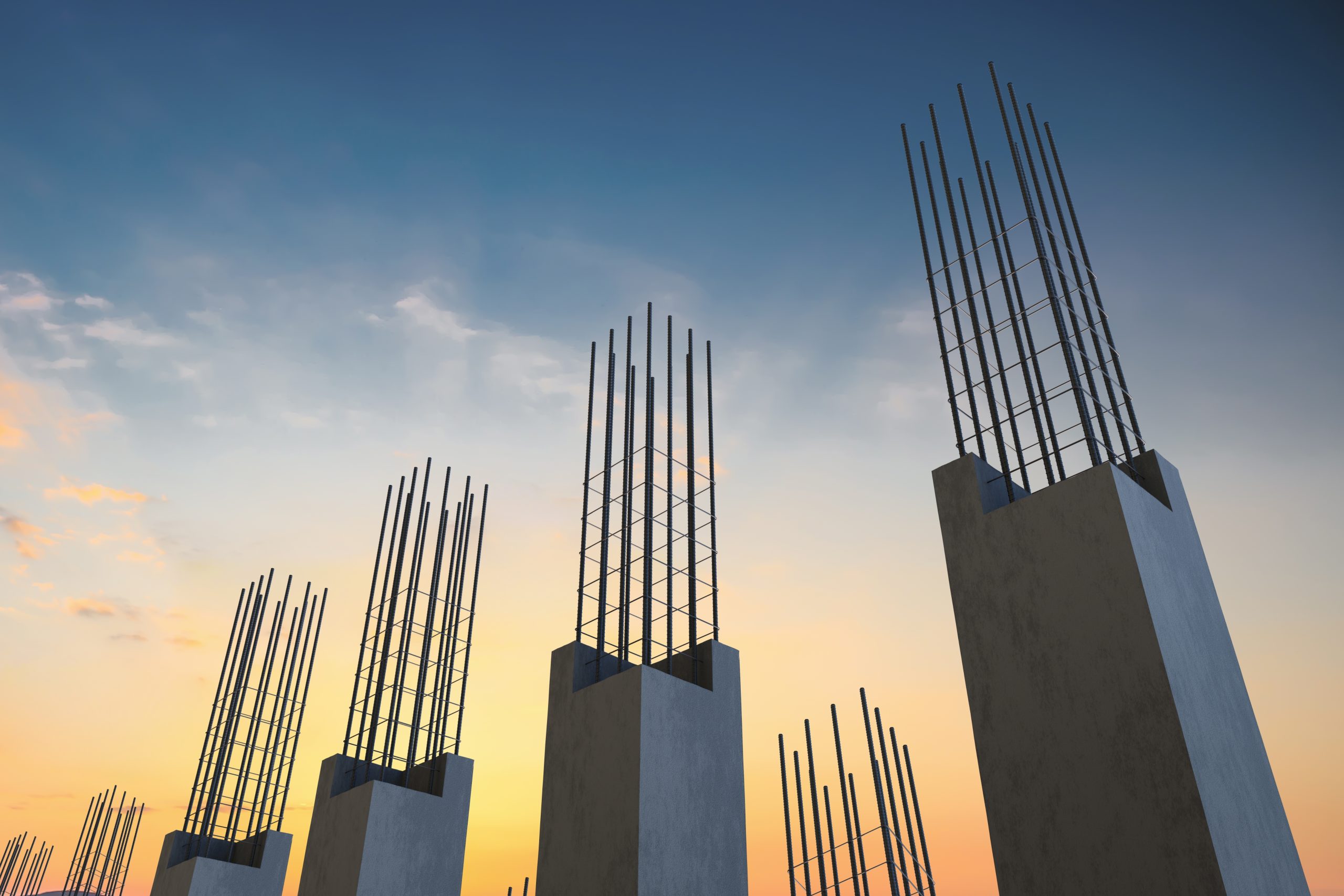 Concrete columns with exposed steel reinforcement bars rise upward at a construction site, silhouetted against a colorful sky during sunset or sunrise.