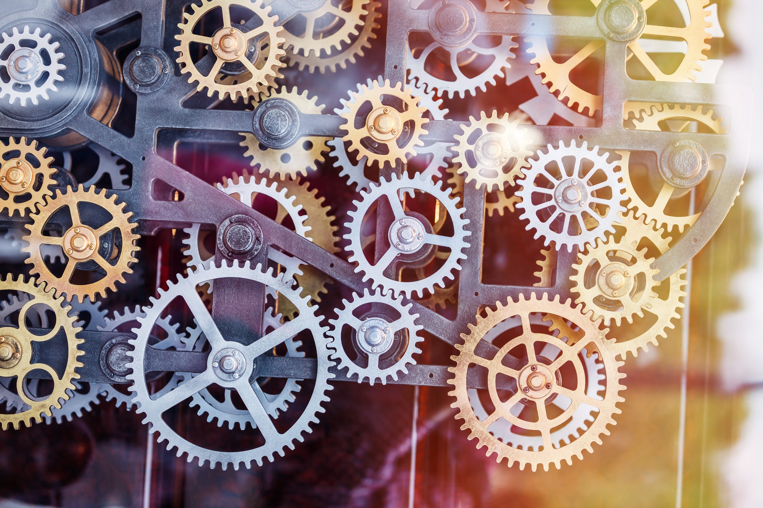 A close-up view of interlocking metal gears and cogs in various sizes, arranged in a complex mechanical system with bright and colorful light reflections.