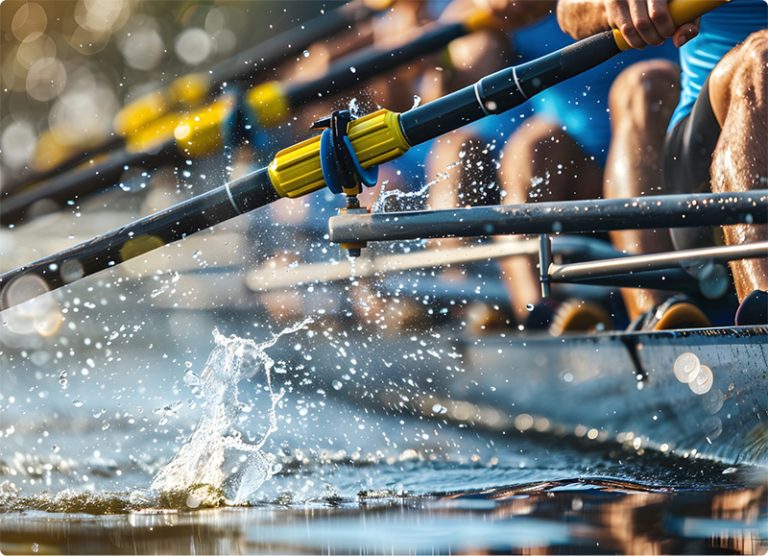 Close-up of several rowers paddling in unison on a racing shell, with water splashing around their oars and blurred, sunlit background.