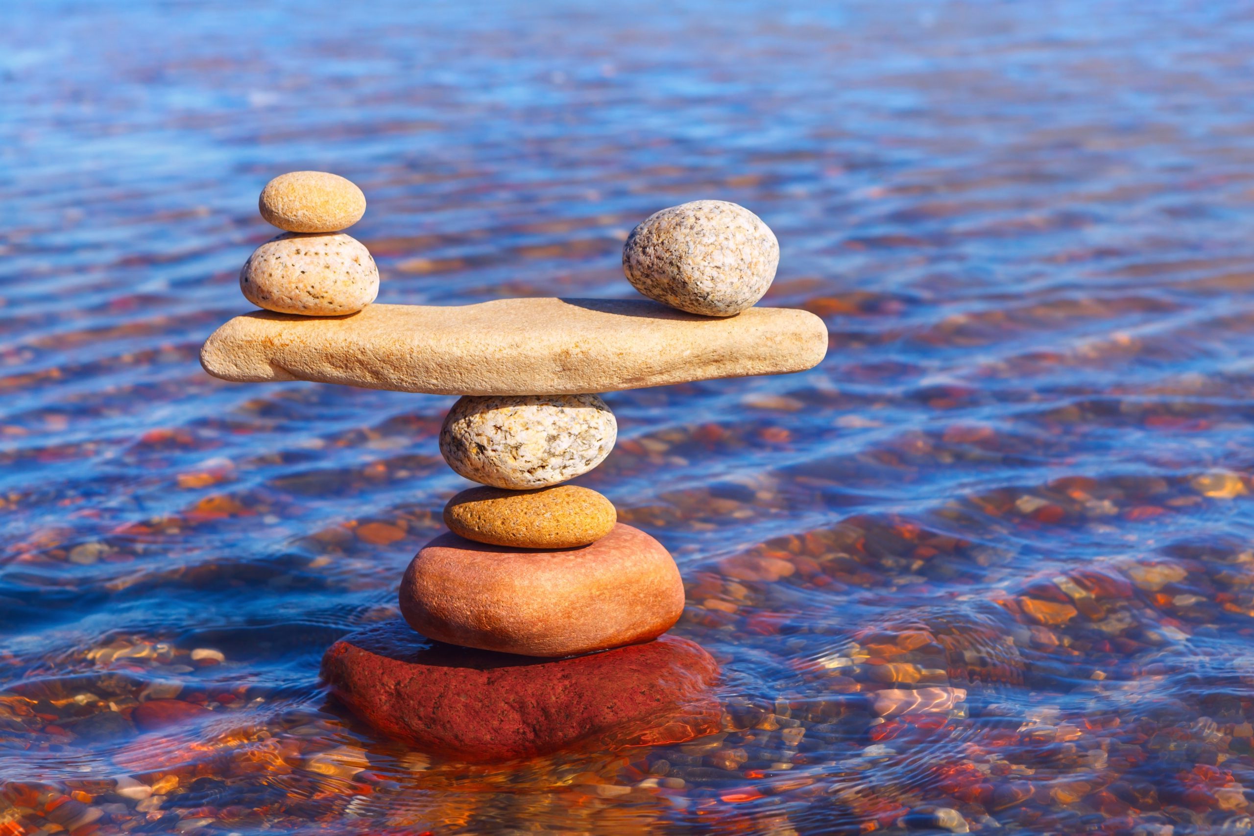 A stack of colorful stones balanced carefully on top of each other in shallow water, with a flat stone in the middle holding two small stones on each end. The water is clear with various pebbles visible beneath the surface.