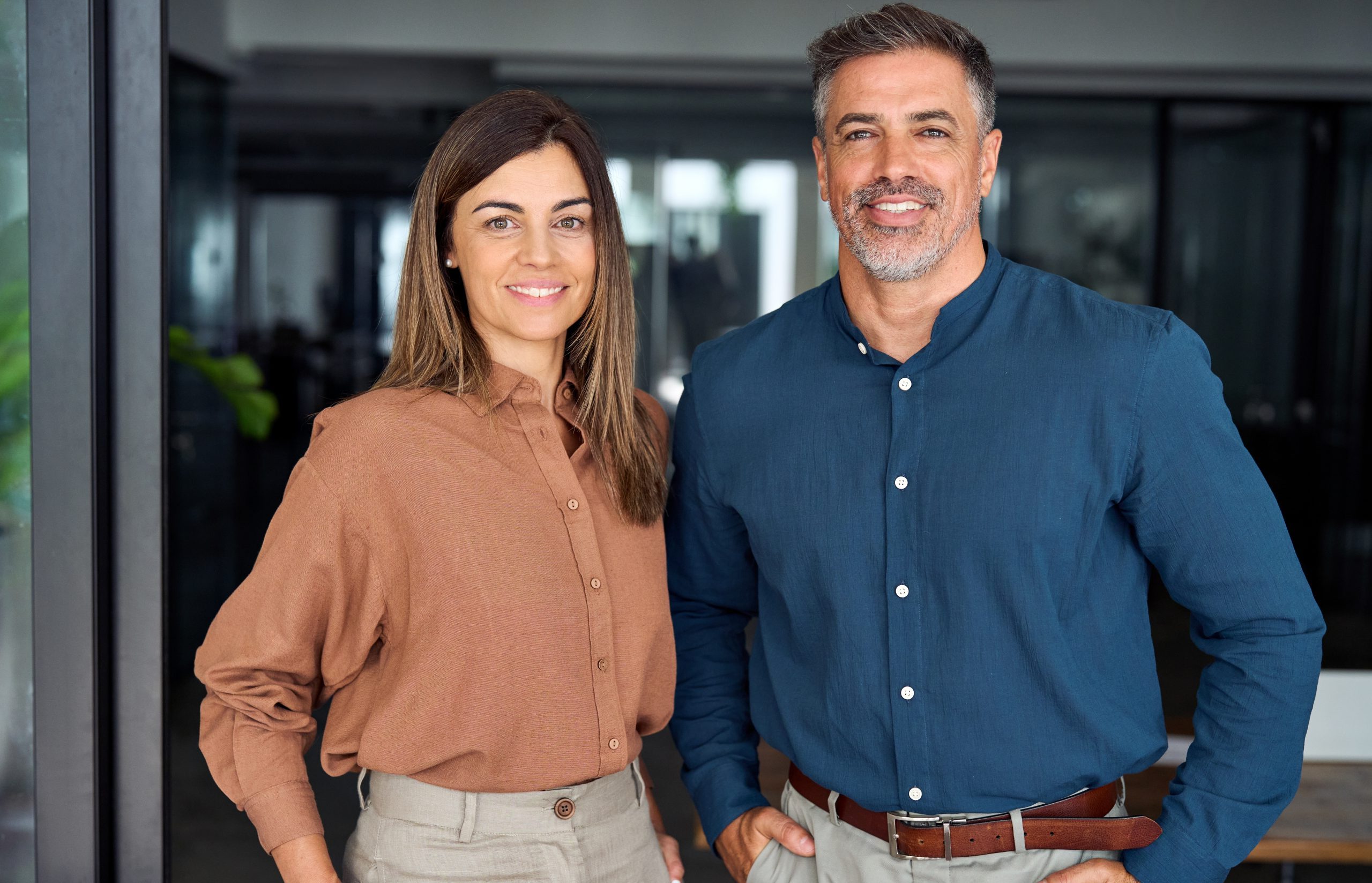 A woman and a man stand side by side indoors, both smiling at the camera. The woman wears a brown shirt and beige pants, while the man wears a blue shirt and gray pants. They appear to be in an office setting.