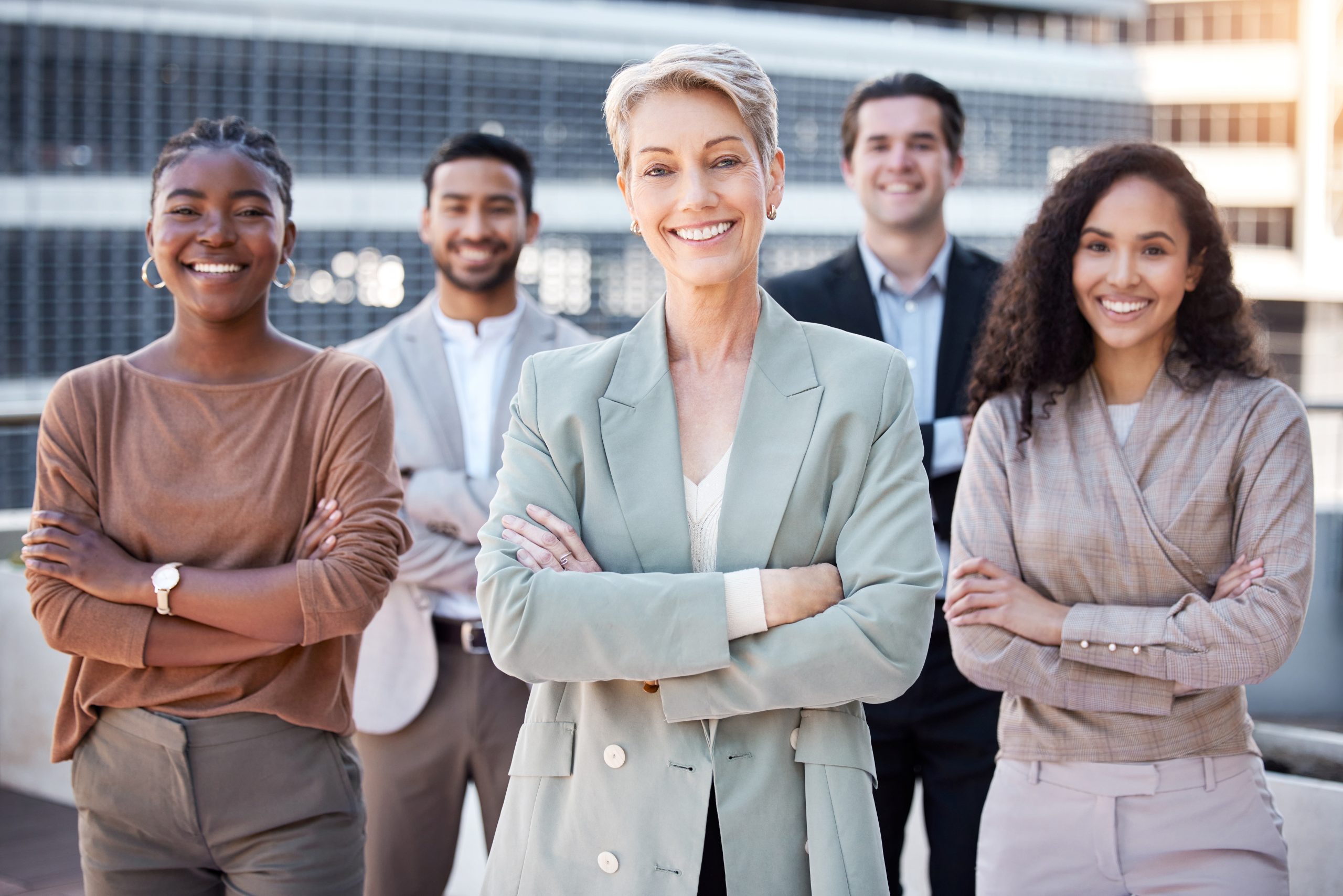 A diverse group of five smiling business professionals standing outdoors with arms crossed, representing teamwork, leadership, and corporate success.