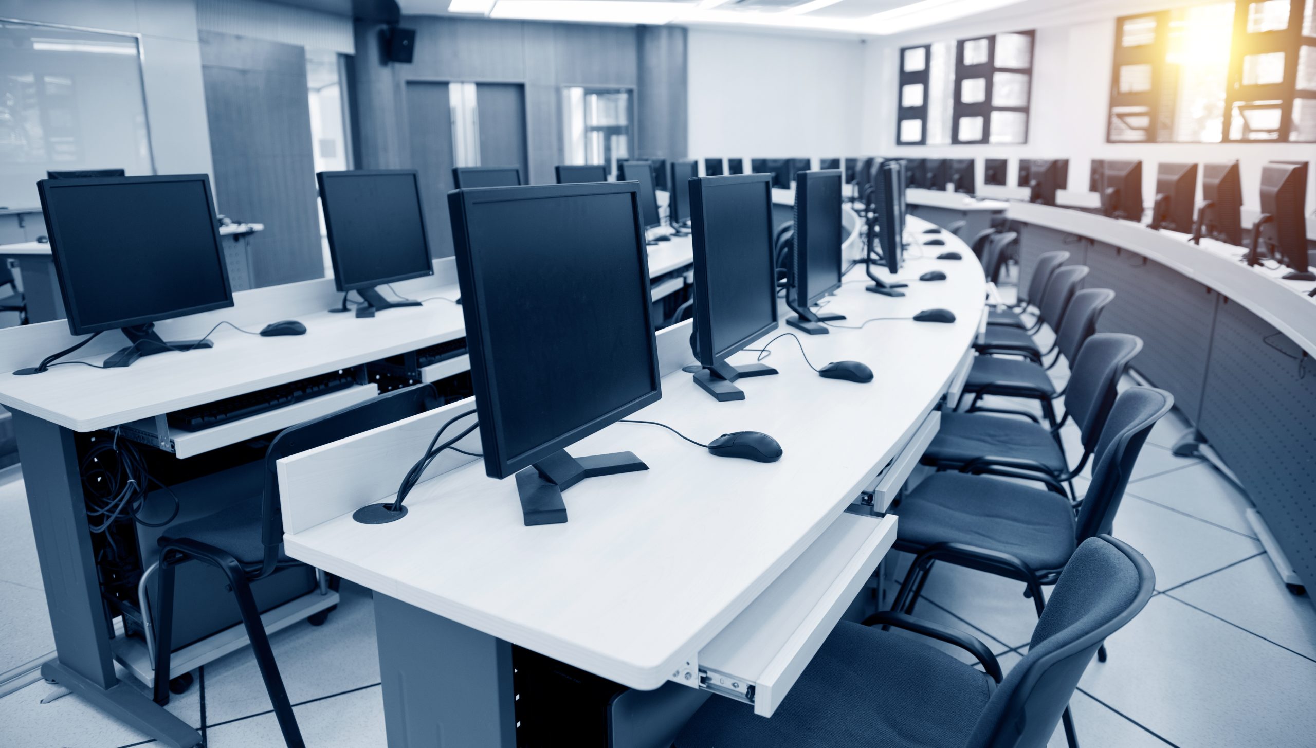 A modern computer lab with rows of desks, each equipped with a monitor, keyboard, and mouse. The room is empty, and sunlight streams through the windows, creating a bright and orderly atmosphere.